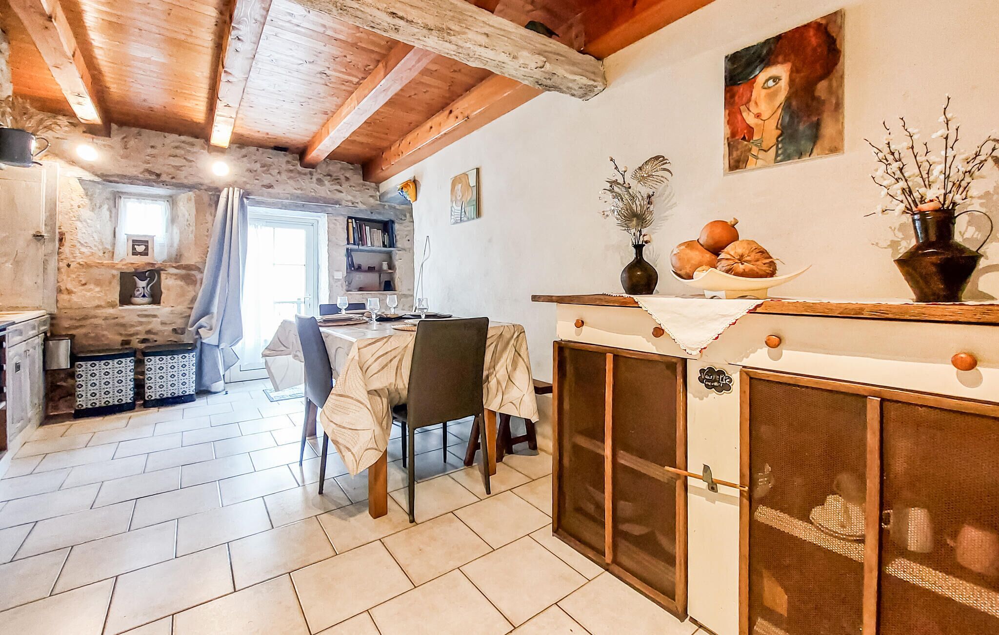 A rustic kitchen with a stone wall, wooden beams, a dining table with chairs, a fireplace, and a bookshelf.