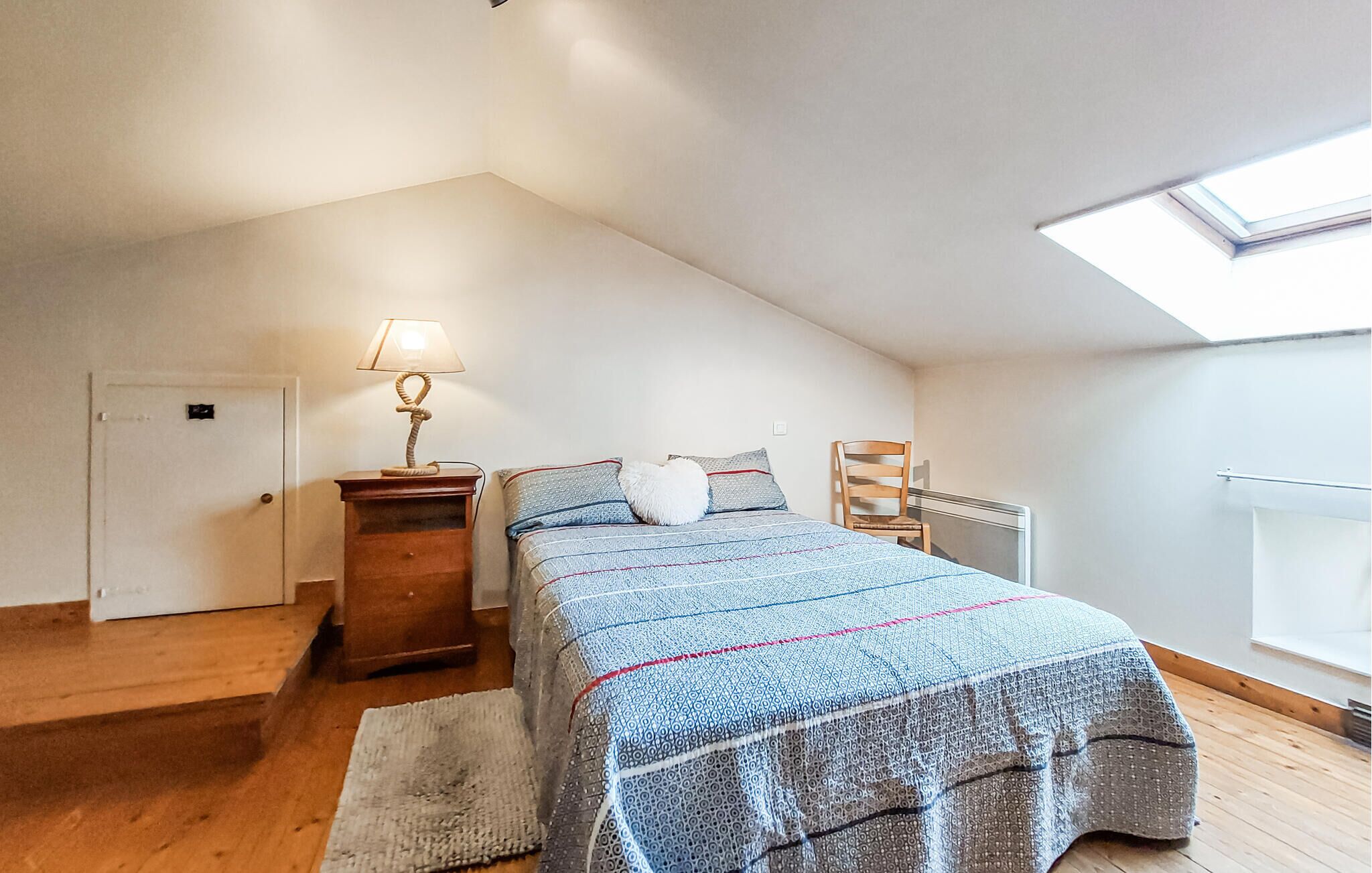 Camera da letto con soffitto spiovente, letto con copriletto decorato, lampada da comodino, una sedia e un termosifone.
