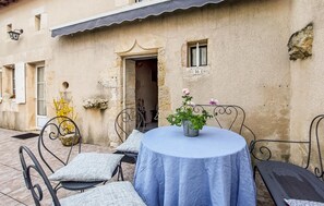 An outdoor patio with a round table and metal chairs, a potted plant, and a building with a stone facade. - Charmante maison au calme (Savigné)