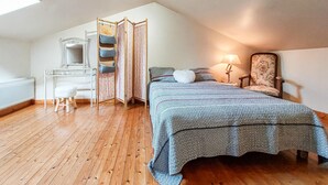 A bedroom with a sloped ceiling, a bed with patterned bedding, a bedside table with a lamp, a chair, and a wardrobe with hanging storage. - Charmante maison au calme (Savigné)