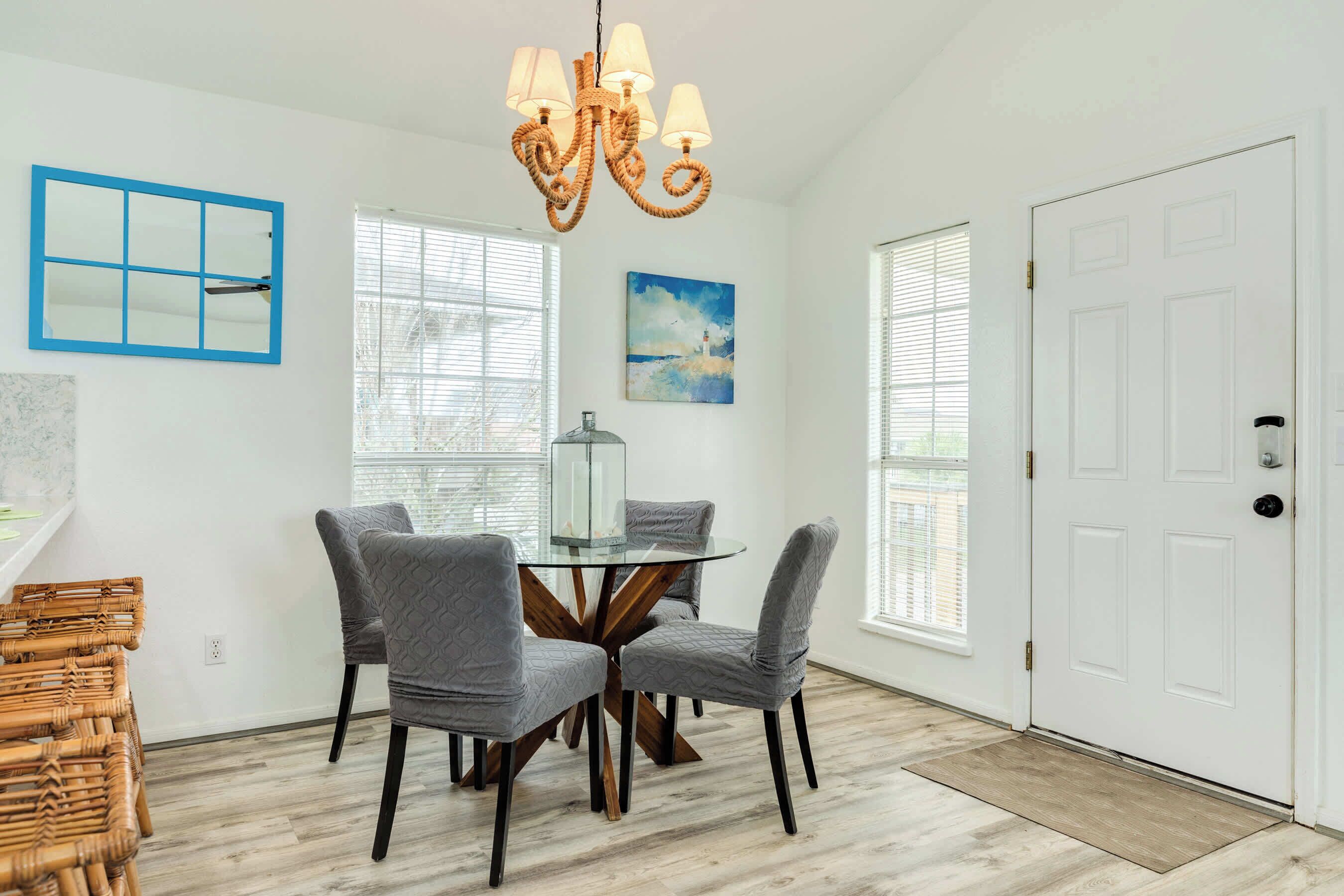 A dining area with a round glass table, four grey upholstered chairs, a chandelier, and a white door.