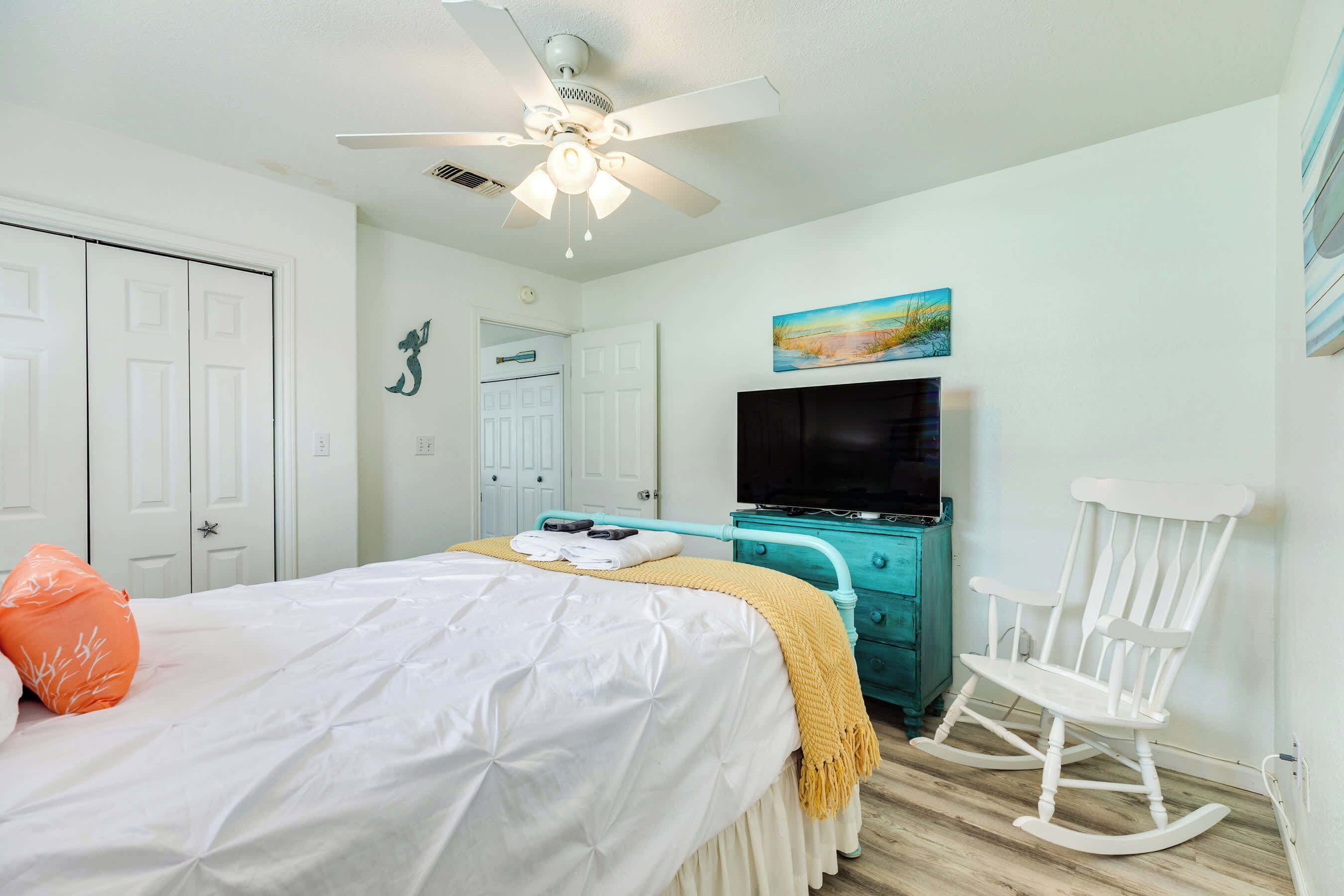A bedroom with a bed, a white rocking chair, a teal dresser, a ceiling fan, and a painting on the wall.