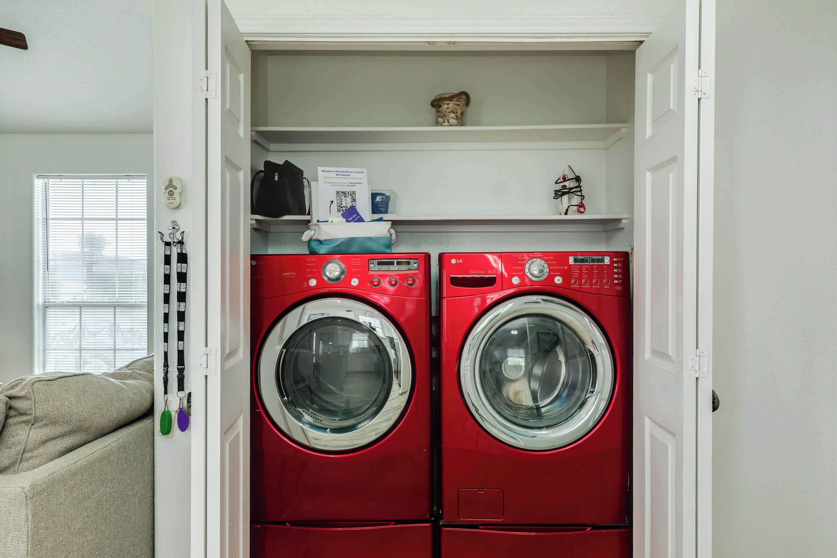 Une buanderie équipée de deux machines à laver et de deux sèche-linge à chargement frontal, tous deux de couleur rouge, une étagère avec une petite figurine et une boîte, et une fenêtre avec des stores.