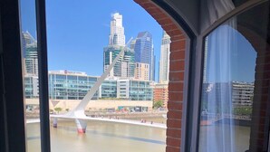 A room with a large window overlooking a cityscape, including a bridge and modern buildings. - Puente de la Mujer, your private apartment with the best view in Bs. As. (Buenos Aires)