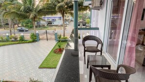 A balcony with wicker chairs and a table, overlooking a street with palm trees and a building in the background. - The Lobos (Benaulim)
