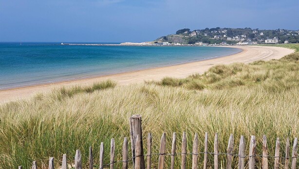 Una playa costera con una valla de madera en primer plano, un mar azul transparente y un pueblo con casas al fondo.
