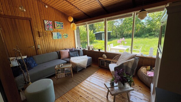 Ett mysigt rum med ett träpanelat tak, en soffa, ett litet bord och utsikt över en trädgård genom stora fönster.