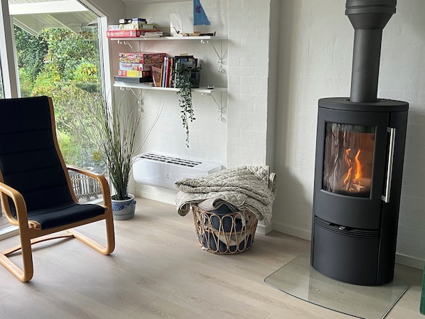 A room with a black wood-burning stove, a chair, a potted plant, a basket, and a shelf with books and boxes. - Lovely holiday home 350 meters from Høll Strand (Børkop)