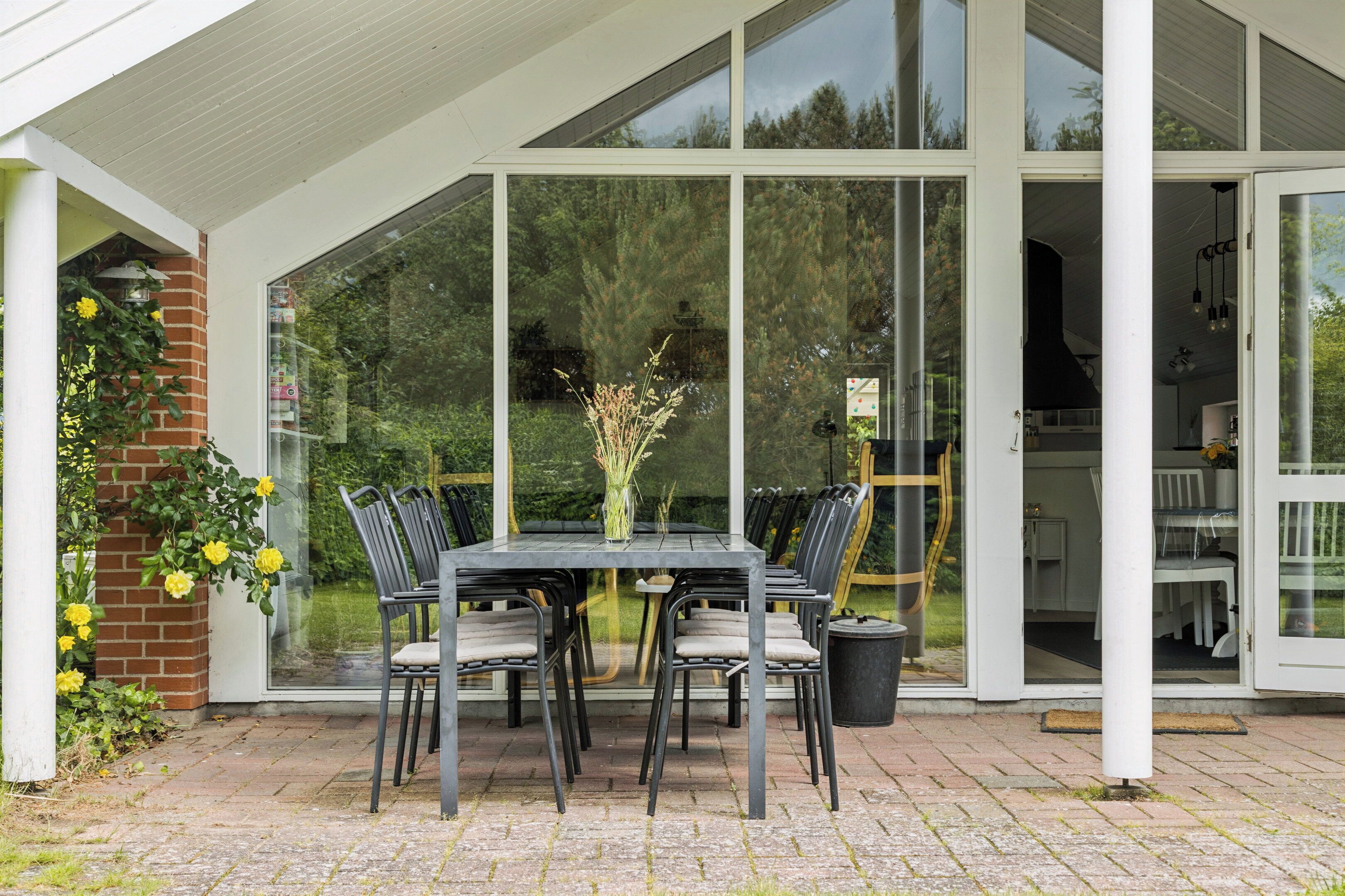 Une terrasse couverte avec une table et des chaises, un mur de briques orné de plantes grimpantes et, à travers de grandes portes vitrées, une vue sur l’intérieur d’une maison.