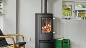 Ein modernes Interieur mit einem schwarzen Kaminofen, einer grünen Kiste mit Holz und einem Sessel.