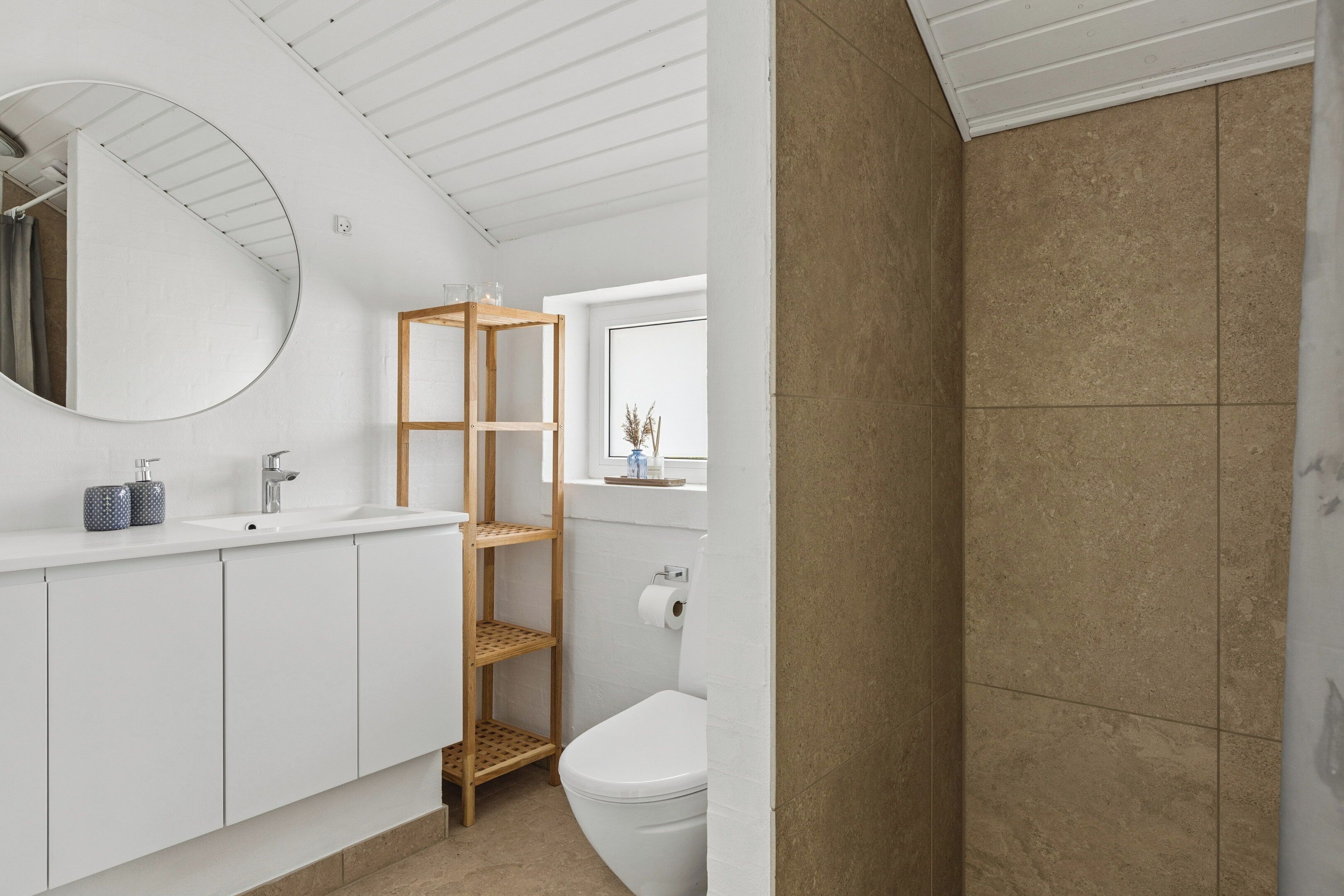 Ein modernes Badezimmer mit einem weißen Waschtisch, einem hölzernen Regal, einem runden Spiegel und einem duschgeflasterten Bereich in Beige.