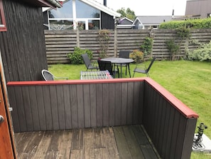 Una terraza de madera con un borde rojo, una mesa con sillas y un jardín al fondo.