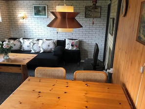 Una habitación con una mesa de madera, un sofá negro con cojines estampados, una lámpara colgante y una pared de ladrillo.