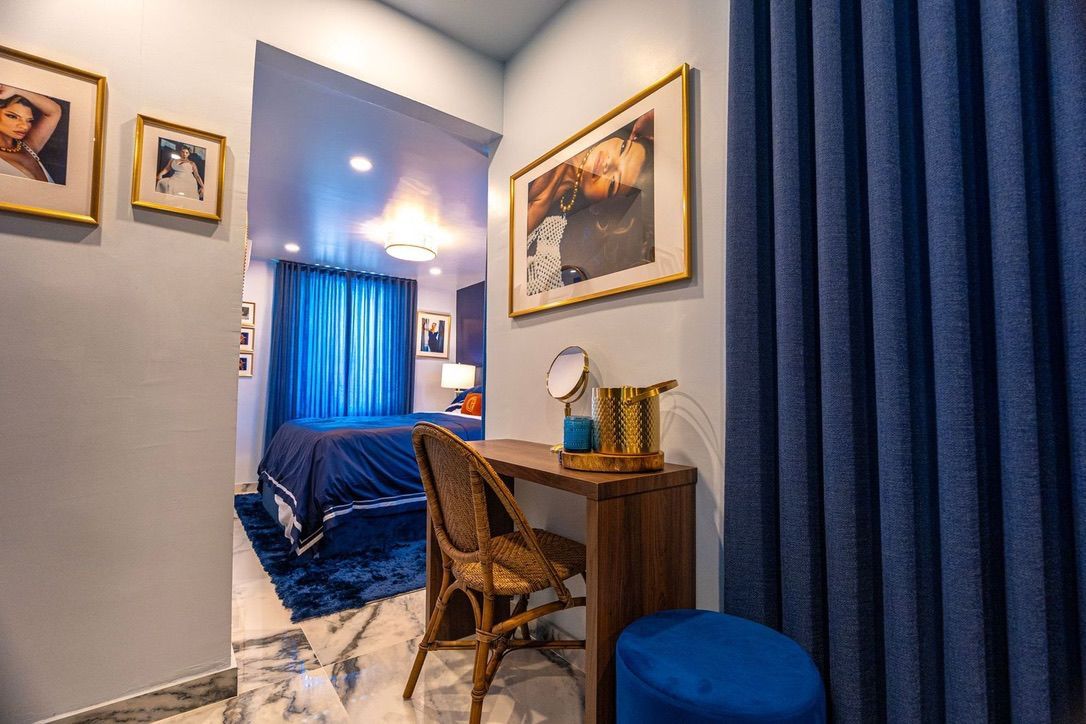 A bedroom with a bed, a desk, a chair, and a bedside table with a lamp and a mirror.