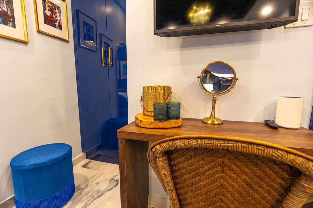 Una habitación con un televisor montado en la pared, un espejo redondo, un taburete azul, una silla de mimbre y una mesa de madera con objetos decorativos.