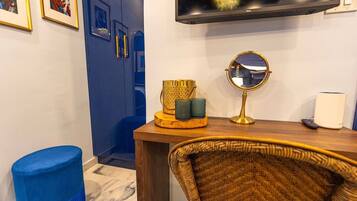 Una habitación con un televisor montado en la pared, un espejo redondo, un taburete azul, una silla de mimbre y una mesa de madera con objetos decorativos.