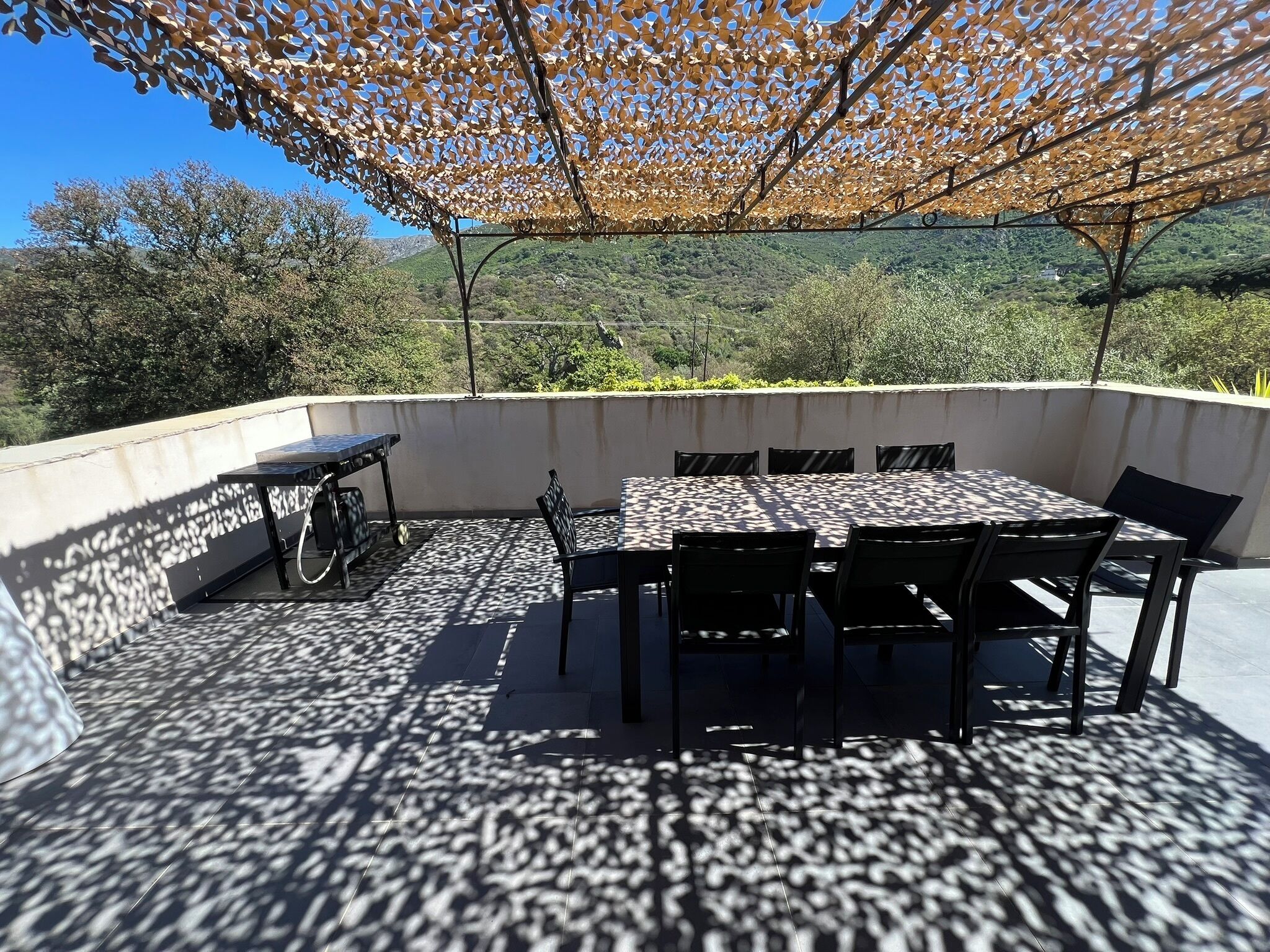 Um pátio externo com mesa e cadeiras, uma bicicleta e uma pérgula que oferece sombra.