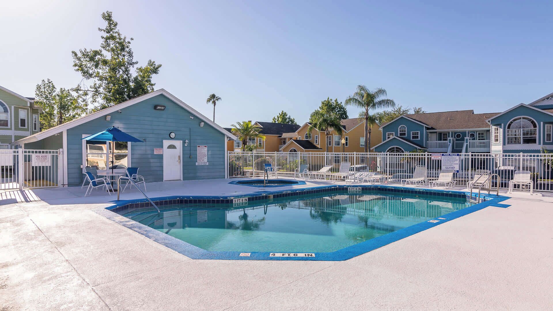 A pool area with a small building, lounge chairs, and a fenced pool.