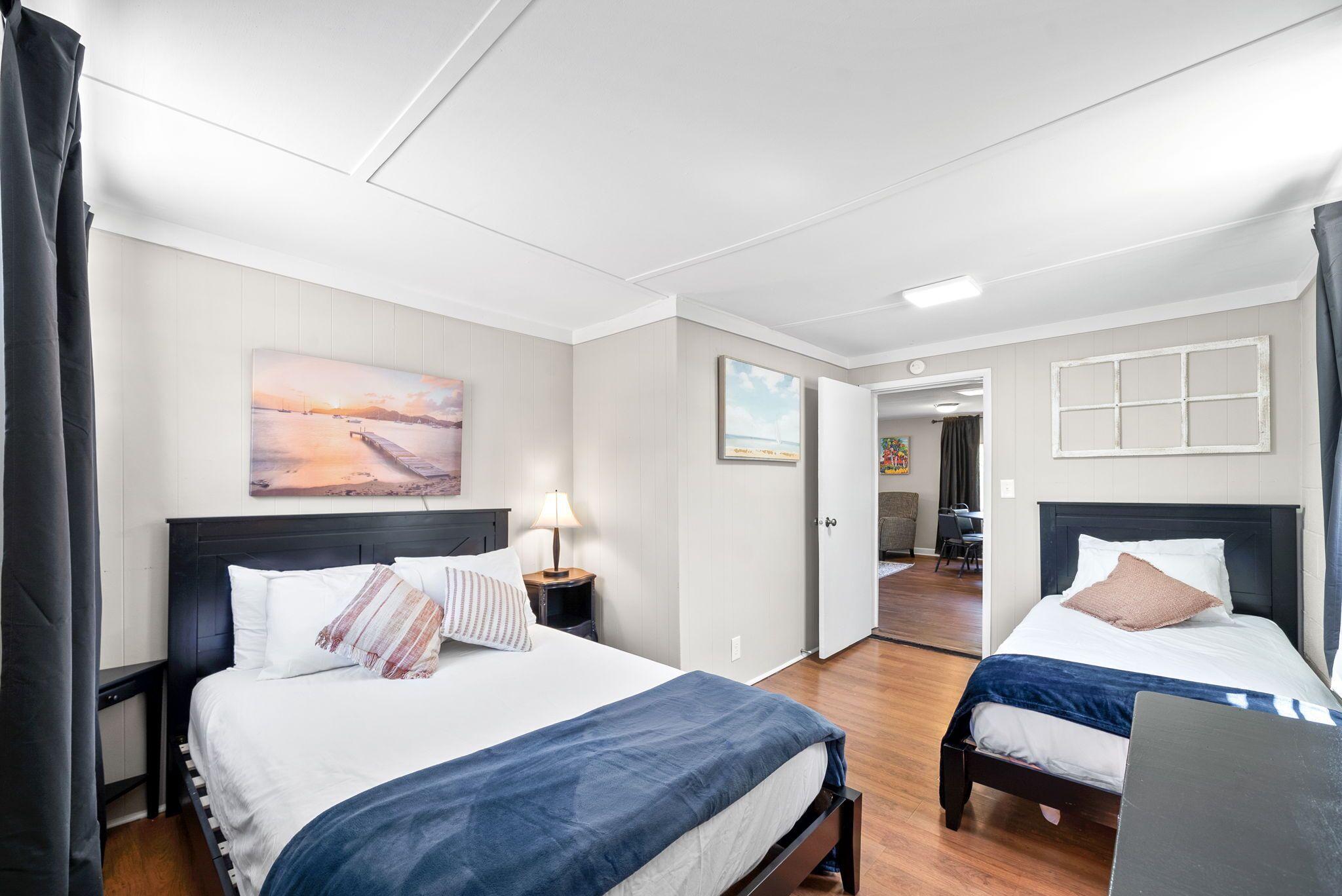 Une chambre d’hôtel avec deux lits, une table de chevet avec une lampe, un tableau au mur et une vue sur le couloir.