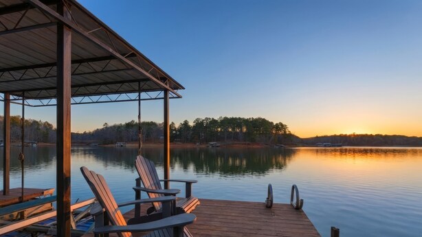 Un muelle de madera con sillas y un área de descanso techada, con vista a un lago tranquilo al atardecer.