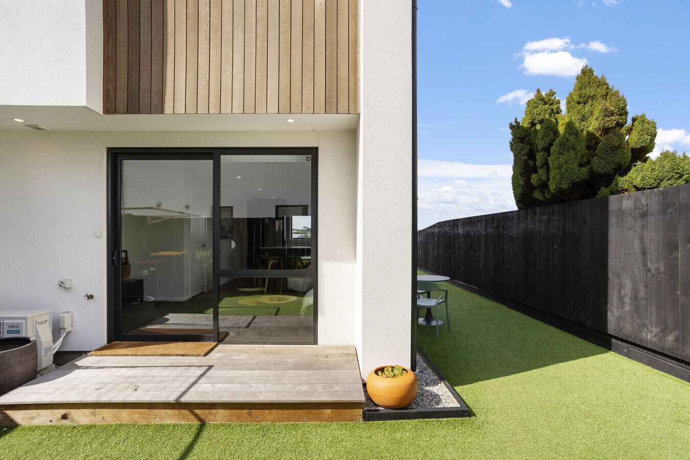 A modern house with a glass door entrance, a small patio with a table and chairs, and a well-maintained lawn.