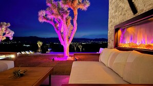 Ein gemütlicher Außenbereich mit einer Couch und einem Holztisch, ein dekorativer Baum, beleuchtet mit violetten Lichtern, und ein Kamin an der Wand.