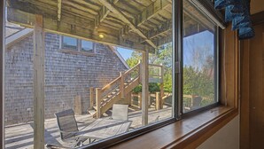 A wooden deck with a staircase leading to a covered area, a window with a view of a building, and outdoor furniture. - Coastal Escape with Fireplace, Three Decks & Easy Beach Access (Cannon Beach)
