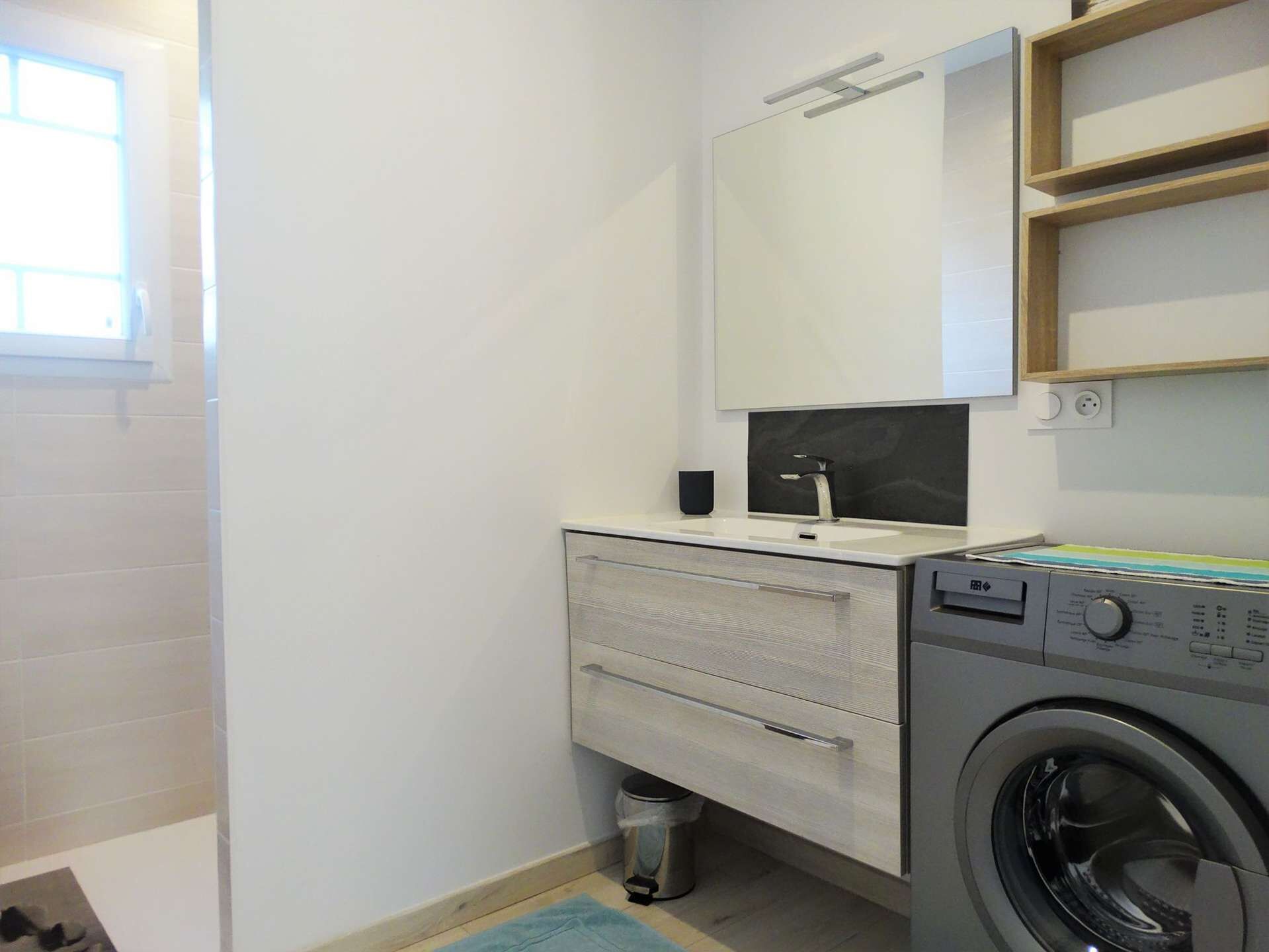 A compact bathroom with a washing machine, a sink with a mirror, and a small trash bin.