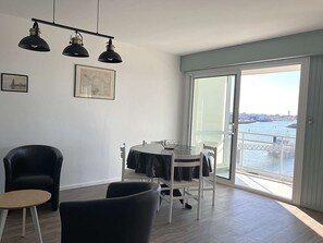 A modern living room with a dining table, chairs, a black leather sofa, a small round table, and a large sliding glass door leading to a balcony with a view of the water and buildings. - Drapeau - Saint Gilles Croix de Vie (SAINT GILLES CROIX DE VIE)