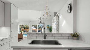 A modern kitchen with white cabinetry, a stainless steel sink, pendant lighting, and a view of the backyard.