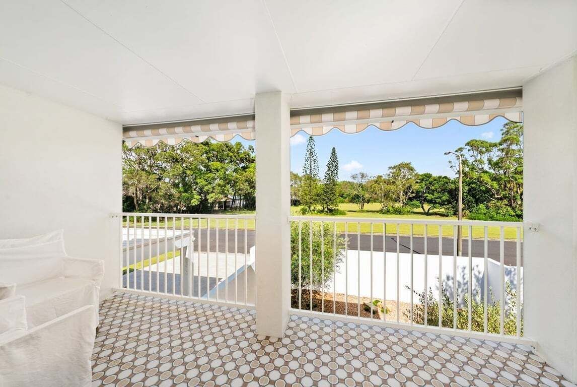 Ein Balkon mit Fliesenboden und weißen Geländern, mit Blick auf einen grünen Park und eine Straße.