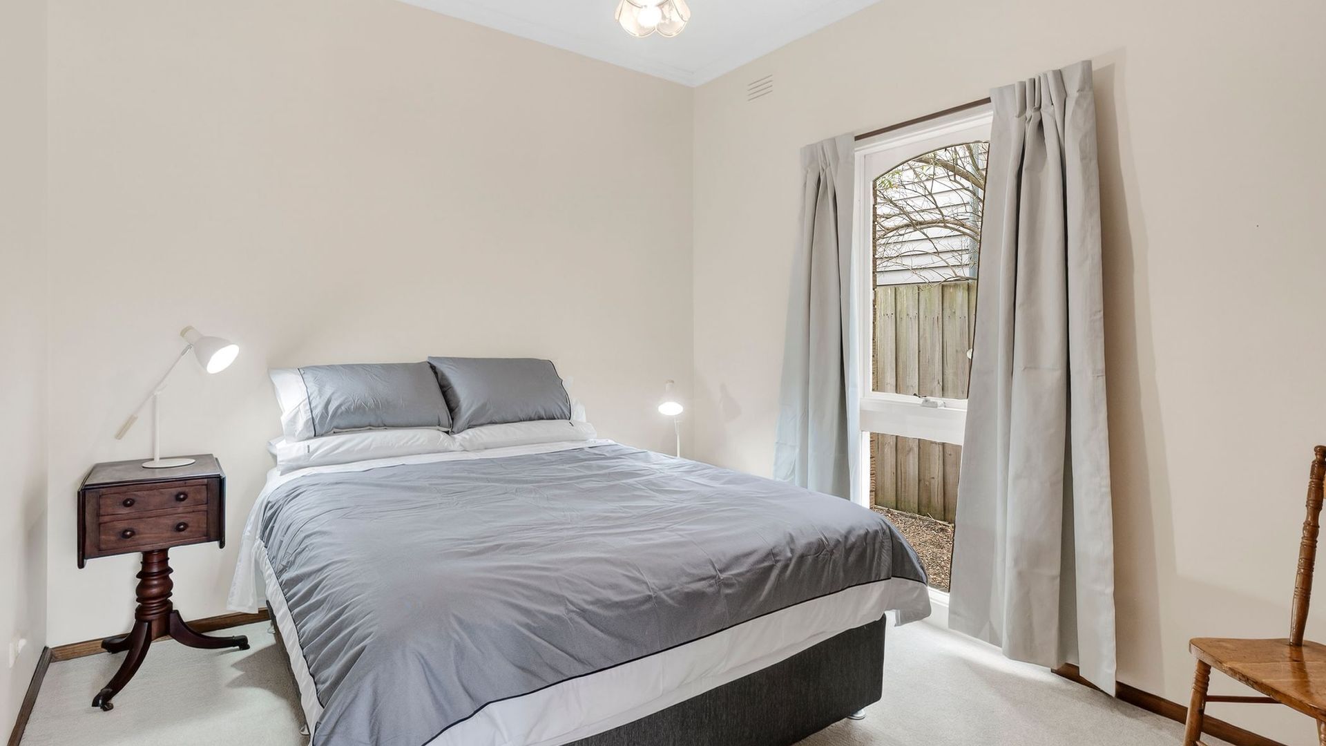 A bedroom with a bed, bedside lamps, a nightstand, a window with curtains, and a view of a wooden fence.