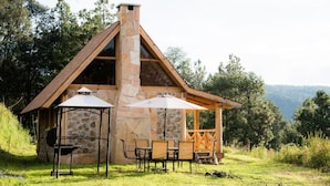 A stone cabin with a chimney, a covered patio with outdoor furniture, and a view of the surrounding forest. - Canto del Bosque (Nanacamilpa)
