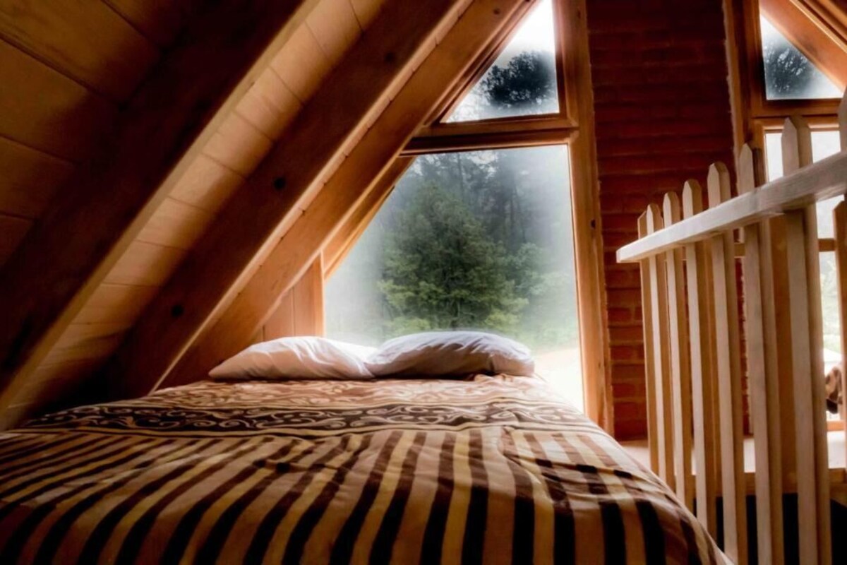 A bedroom with a sloped ceiling, a bed with patterned bedding, and a view of a forest through a window.