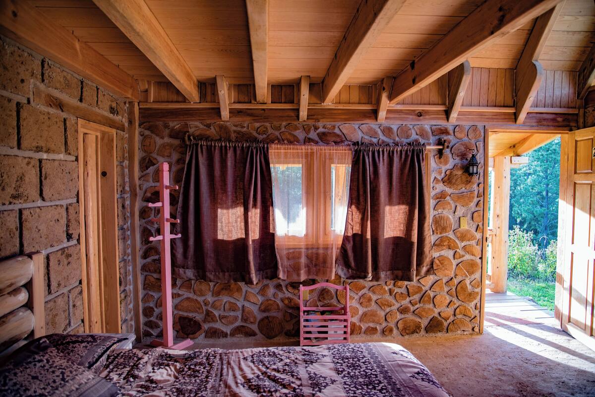 Ein rustikaler Raum mit Steinwänden, Holzbalken, einem Bett mit gemusterten Textilien, einem Holzstuhl und einem Fenster mit Vorhängen.