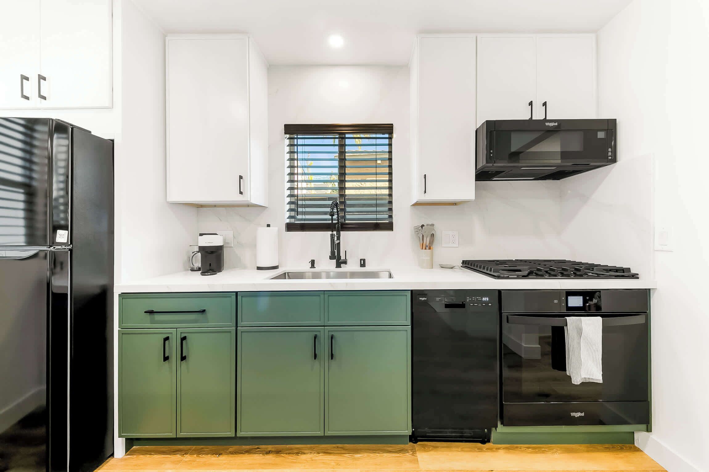 Una cocina moderna con armarios blancos, un microondas negro, un lavavajillas y una ventana con persianas.