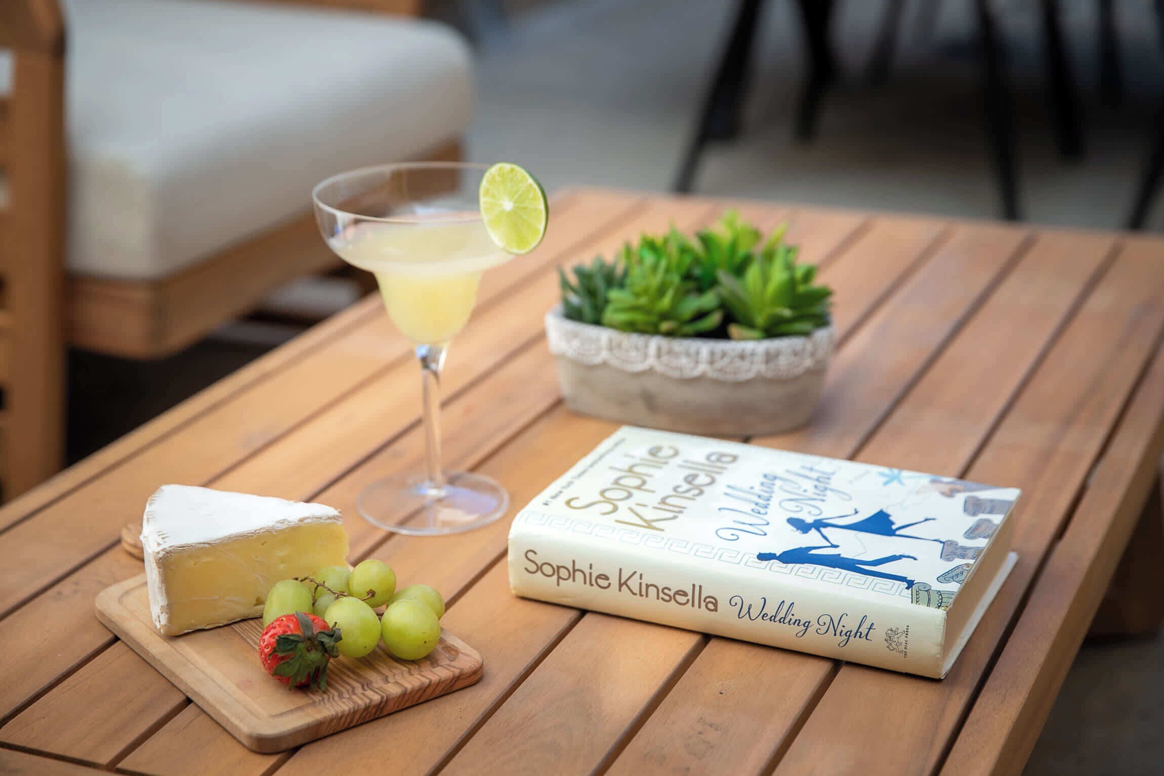 A wooden table with a book titled 'Sophie Kinsella Wedding Night', a glass of cocktail with a lime garnish, a piece of cheese, and grapes.