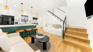 A modern kitchen with a central island, a dining area with bar stools, a living room with a sofa, and a staircase with a black railing. - PB New Build - Steps to Sand & Surf (San Diego)