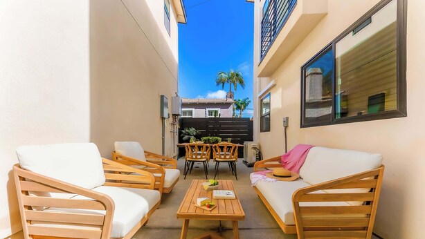 A narrow outdoor patio with wooden furniture, including two sofas and a table, and a view of a building in the background. - PB New Build - Steps to Sand & Surf (San Diego)