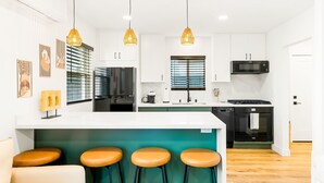 A modern kitchen with white cabinets, a central island with stools, pendant lighting, and a built-in oven. - PB New Build - Steps to Sand & Surf (San Diego)