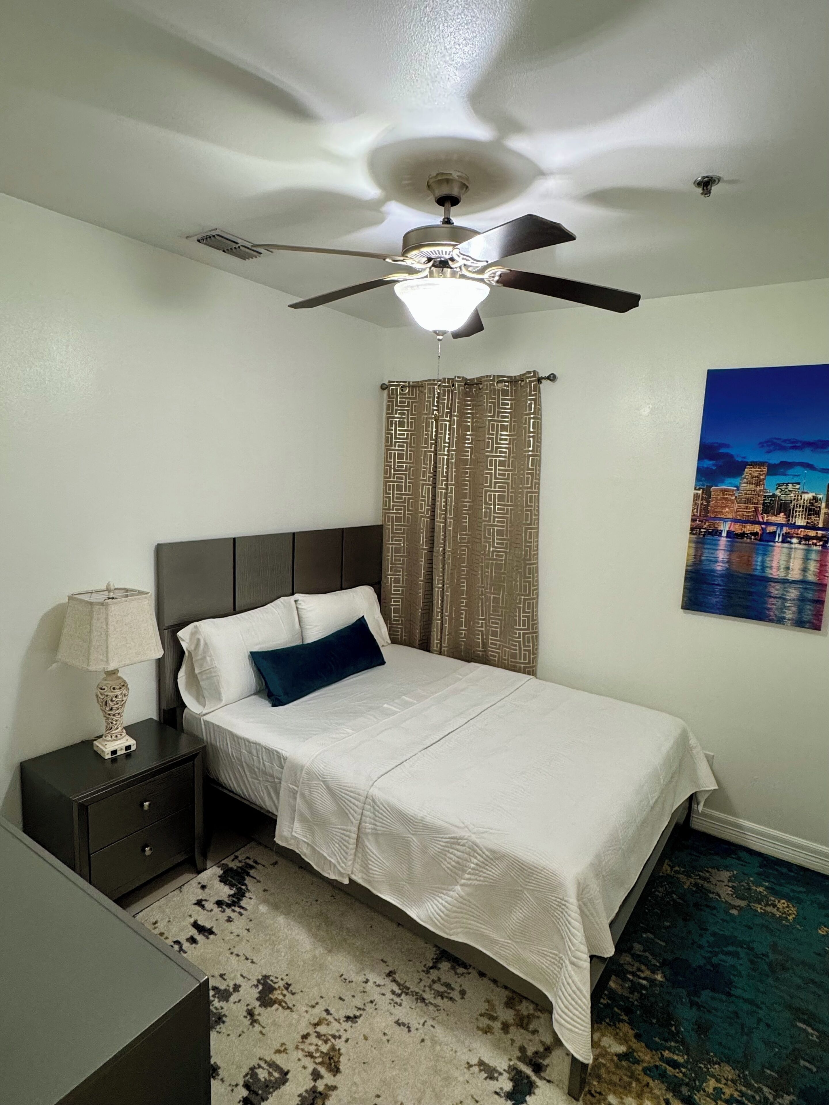 A bedroom with a bed, a nightstand, a ceiling fan, a patterned curtain, and a cityscape painting on the wall.