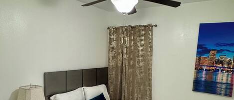 A bedroom with a bed, a nightstand, a ceiling fan, a patterned curtain, and a cityscape painting on the wall.