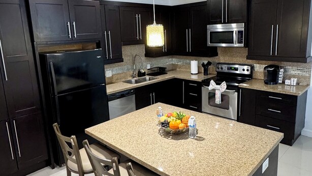 A modern kitchen with dark cabinets, stainless steel appliances, a granite countertop, and a central island with bar stools. - The Prestige 3BR/2BA Place Near Airport and Mall (McAllen)