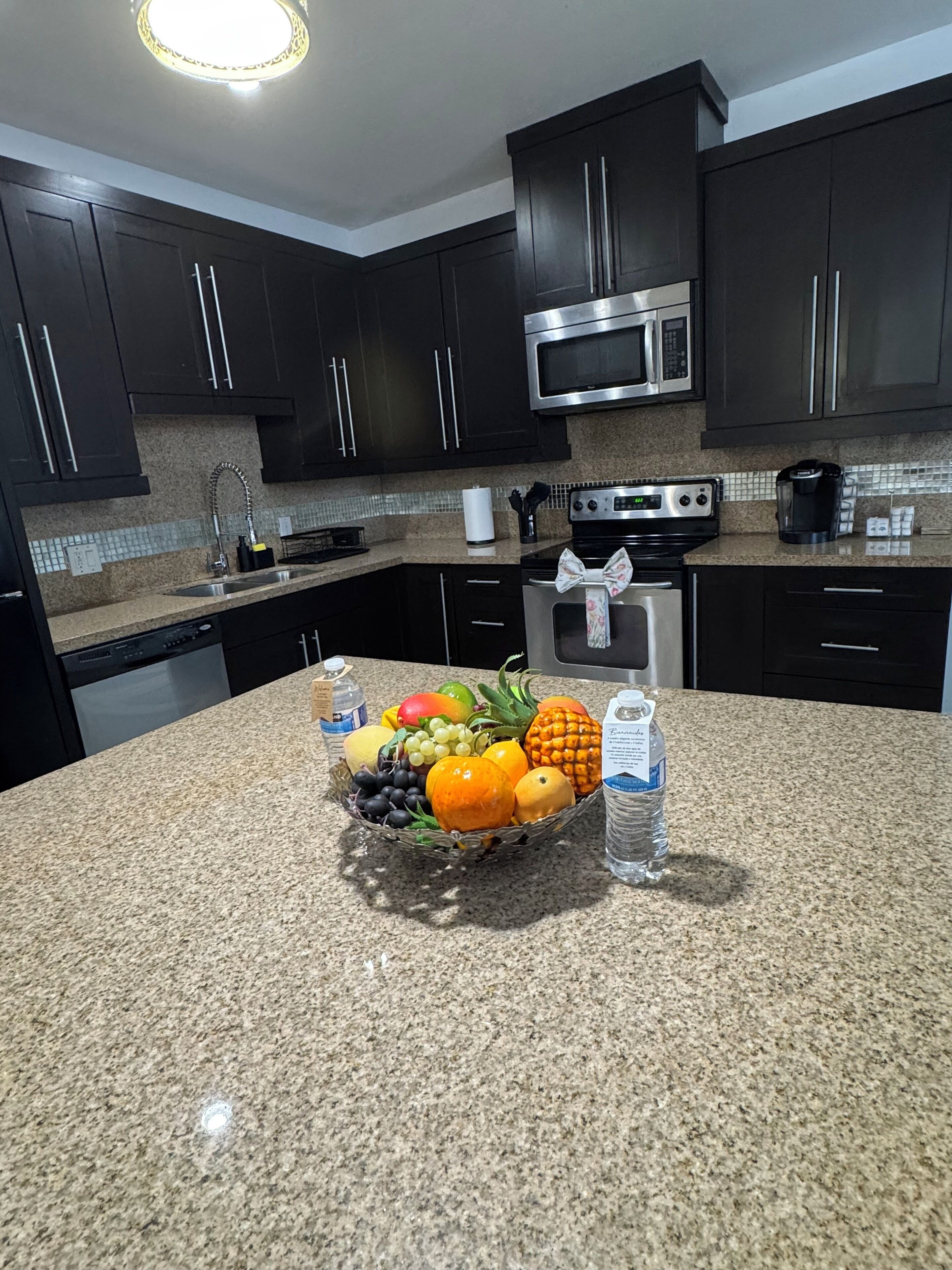 A kitchen with dark cabinets, a stainless steel microwave, a coffee maker, and a fruit basket on the counter.
