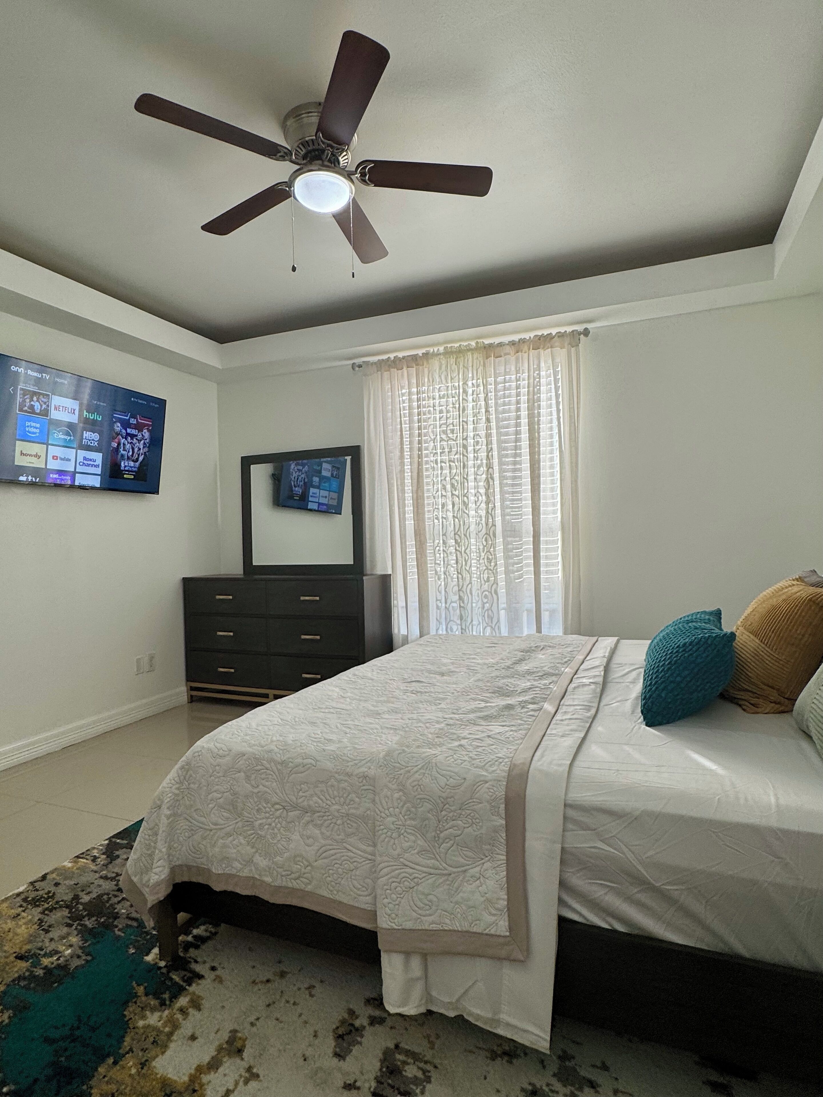 Un dormitorio con cama, ventilador de techo, cómoda, televisor montado en la pared y ventana con cortinas.