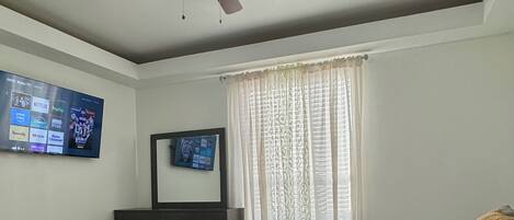 Un dormitorio con cama, ventilador de techo, cómoda, televisor montado en la pared y ventana con cortinas.