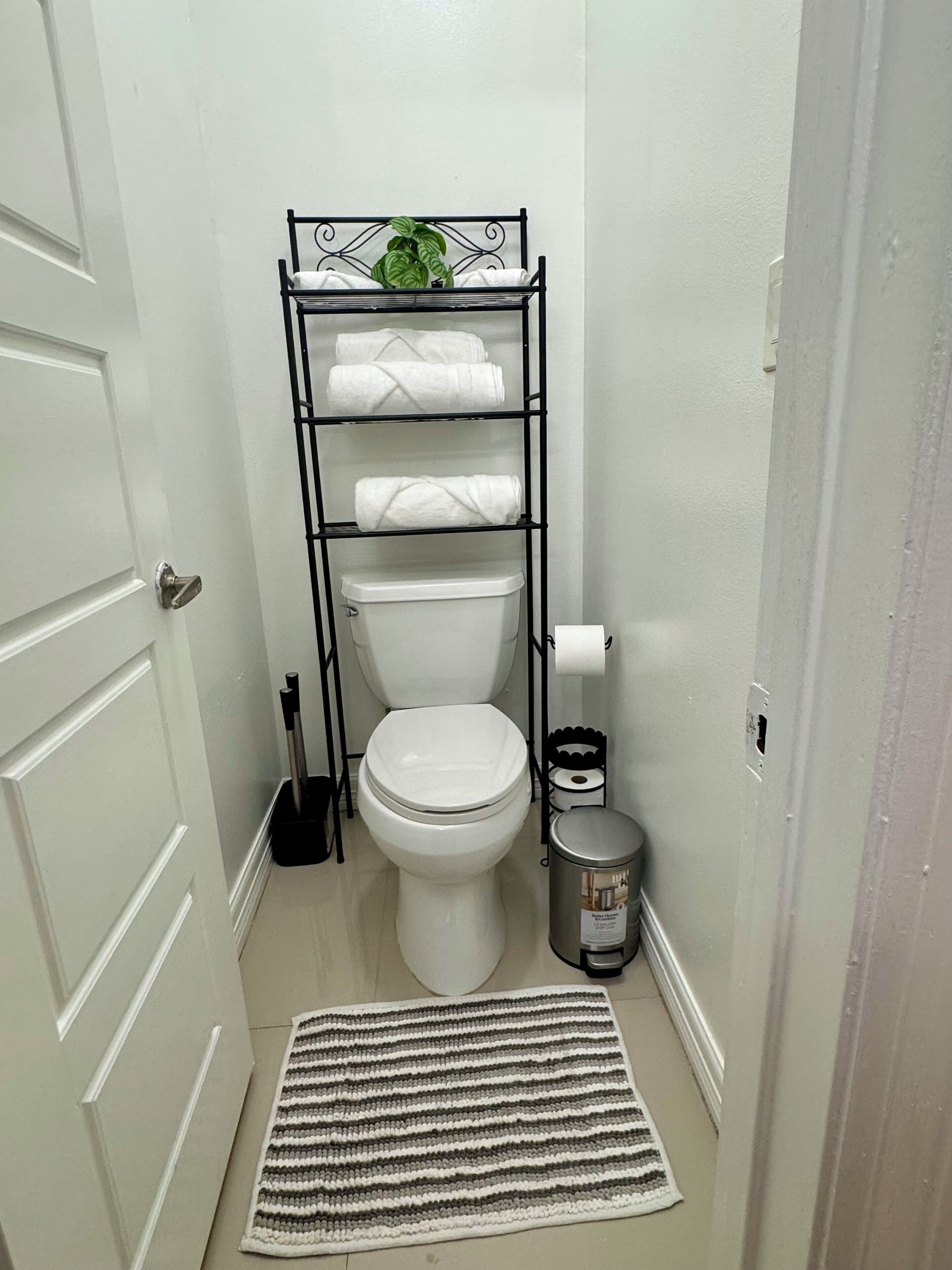 Ein kleines Badezimmer mit Toilette, einem Metallregal mit Handtüchern, einer kleinen Mülltonne und einem Streifen Teppich.