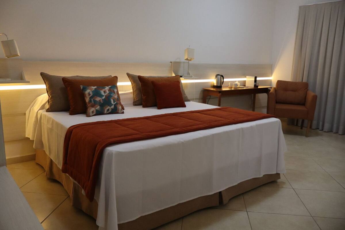 A hotel room with a bed, pillows, a chair, a desk, and a lamp.