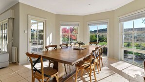 A spacious dining area with a large wooden table and six chairs, large windows offering views of a garden and hills, and a white radiator on the left.