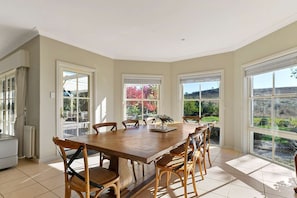 A spacious dining area with a large wooden table and six chairs, large windows offering views of a garden and hills, and a white radiator on the left. - Luxe Countryside Home - Amazing Views, Sleeps 6 (Byng)